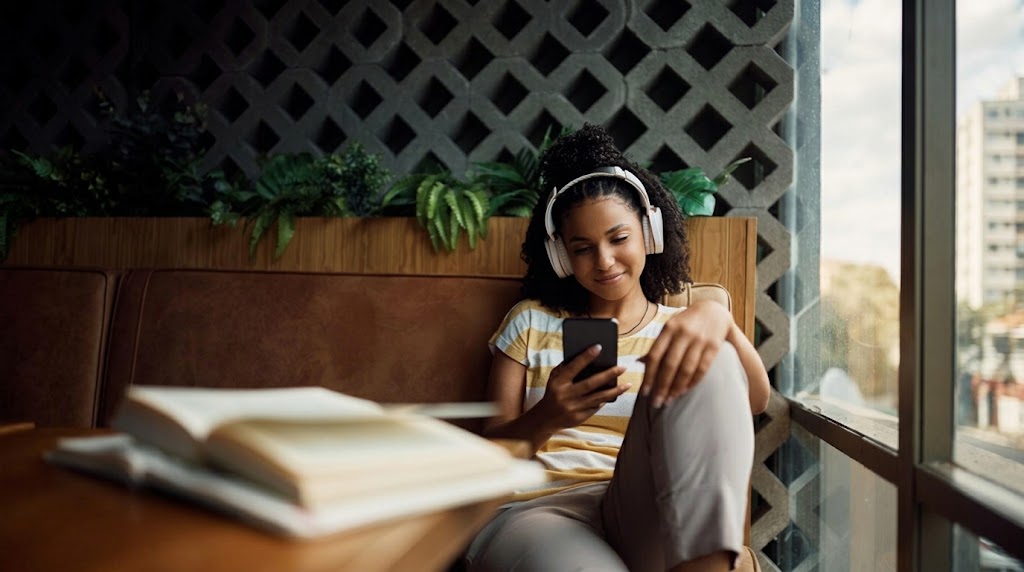 A young woman with headphones listening to her phone while sitting on a brown couch in a cafe