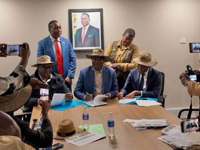 Three seated officials sign documents at a conference table while colleagues and photographers observe the signing ceremony.