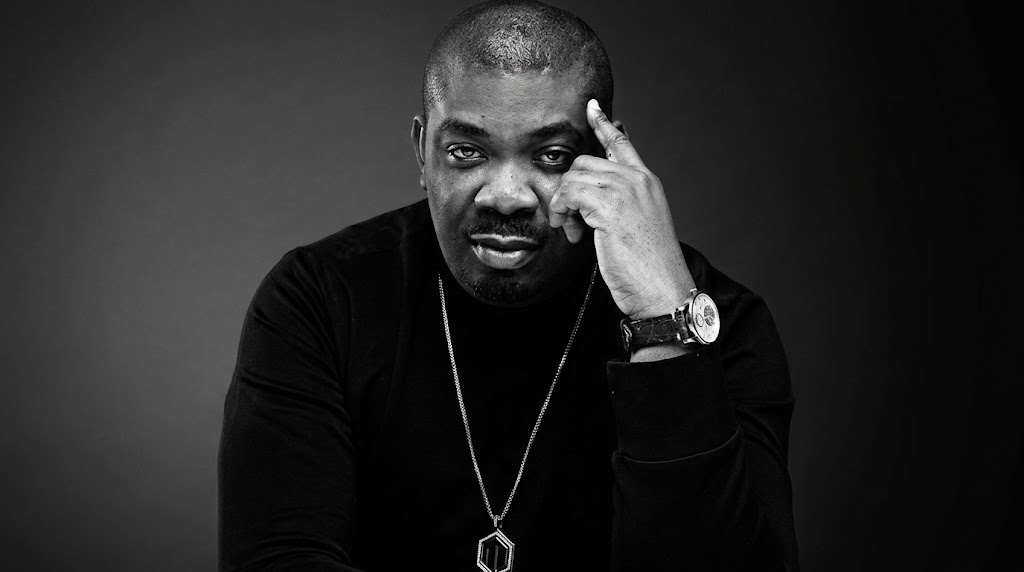 Black and white portrait of Nigerian music producer Don Jazzy wearing a long-sleeved shirt, silver pendant necklace, and wristwatch, posing thoughtfully against a dark studio background.