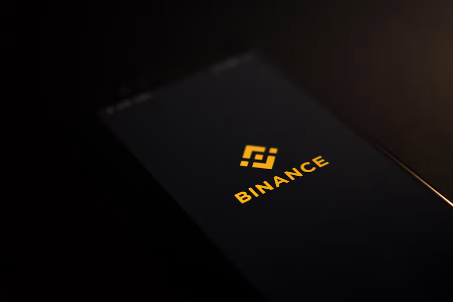 Close-up shot of a smartphone screen displaying the Binance logo in yellow against a dark background, resting on a dark surface.
